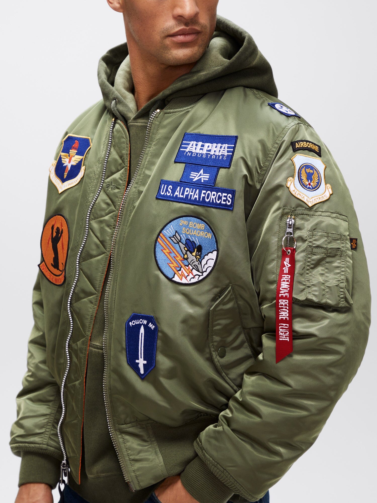 Alpha Industries MA-1 Squadron Bomber Jacket in green with squadron patches and sleeve zip pocket, over a hooded sweatshirt.