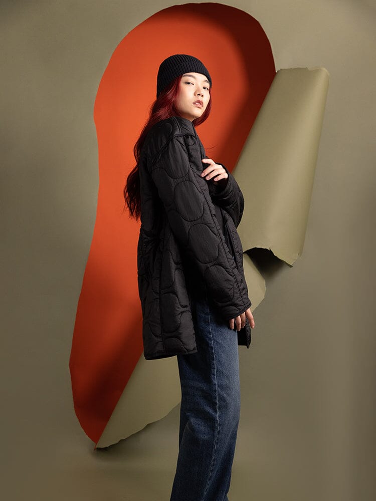 A person wearing a black beanie and the Women's Mixed Media Quilted Long Liner from Alpha Industries, showcasing an onion quilting pattern.