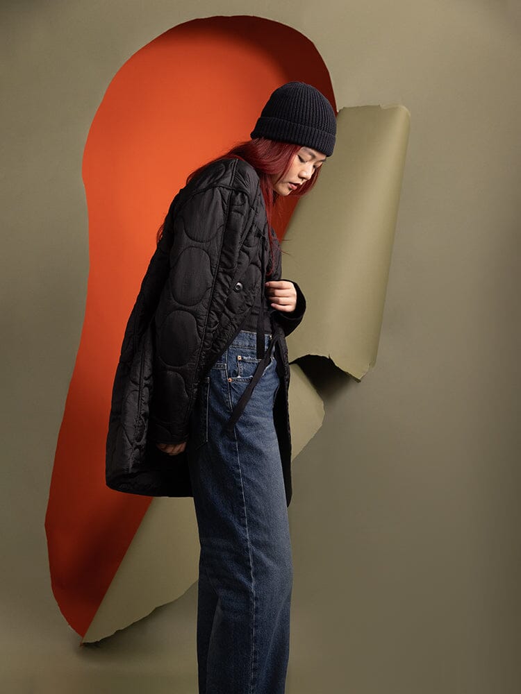 A person wearing the Women's Mixed Media Quilted Long Liner in black by Alpha Industries, paired with a beanie.