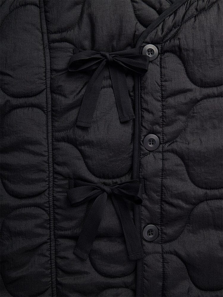 Close-up of the Women's Mixed Media Quilted Long Liner by Alpha Industries, showcasing its black quilted fabric adorned with two black bow ties and three vertically stitched black buttons in an onion quilting pattern.