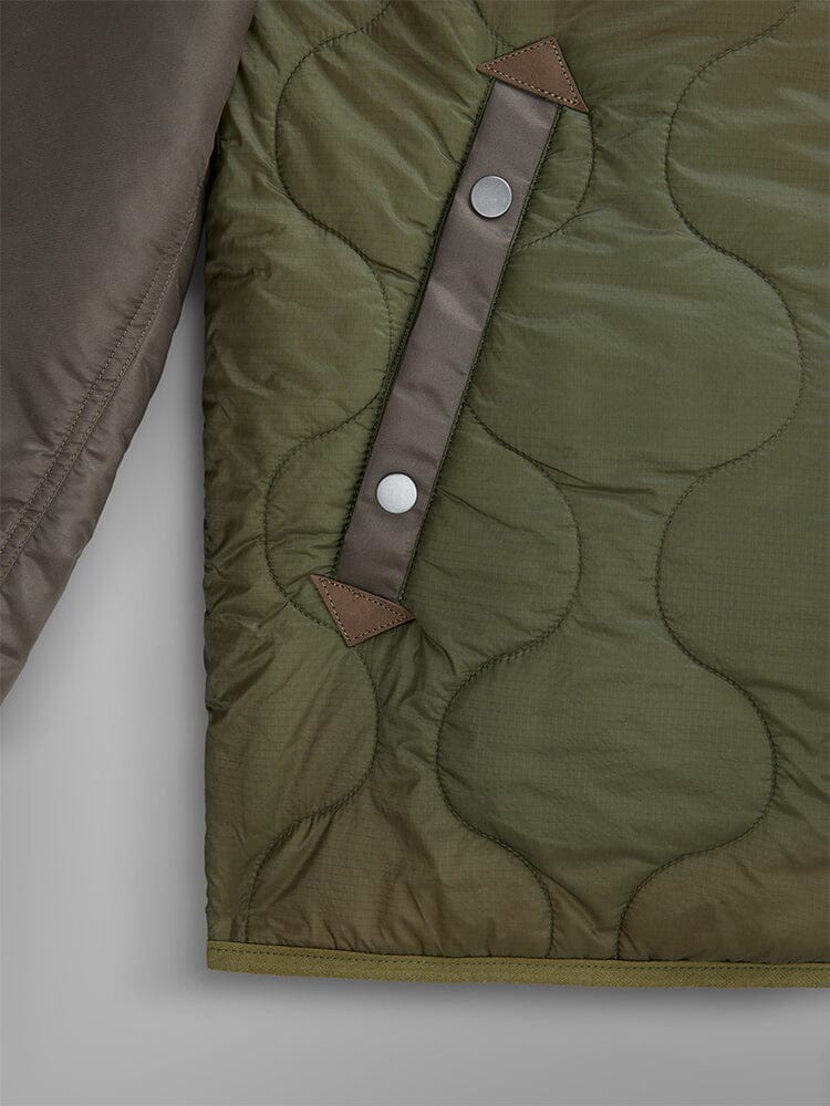 A close-up of the Mixed Media Quilted Utility Jacket by Alpha Industries showcases an onion-quilted olive green design with a wavy pattern, complemented by two silver snap buttons on a vertical strap and brown triangular reinforcements.