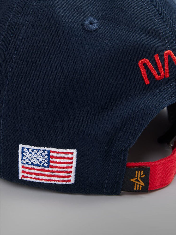 The NASA Worm Logo Cap by Alpha Industries is a navy blue hat adorned with prominent red NASA text and an American flag patch on the side. Shown from the back side.