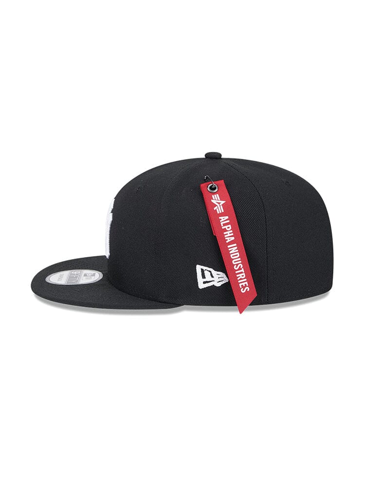Side view of a black New York Yankees x Alpha x New Era 950 Cap. This snapback by Alpha Industries features a red tag and detailed white team logo.