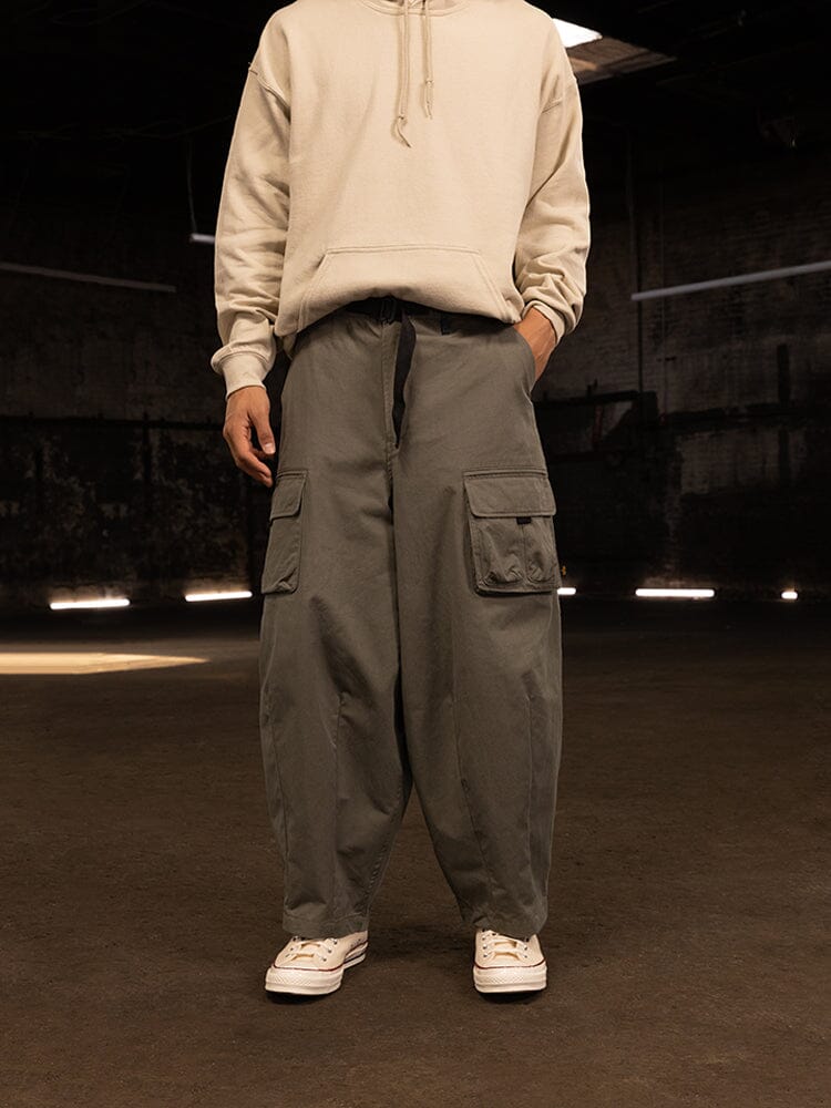 A person wearing a beige hoodie and the Oversized Tactical Pant from Alpha Industries stands indoors, hands tucked casually into the generous cargo pockets of the stylish gray pants with an elastic waistband, paired with classic white sneakers.