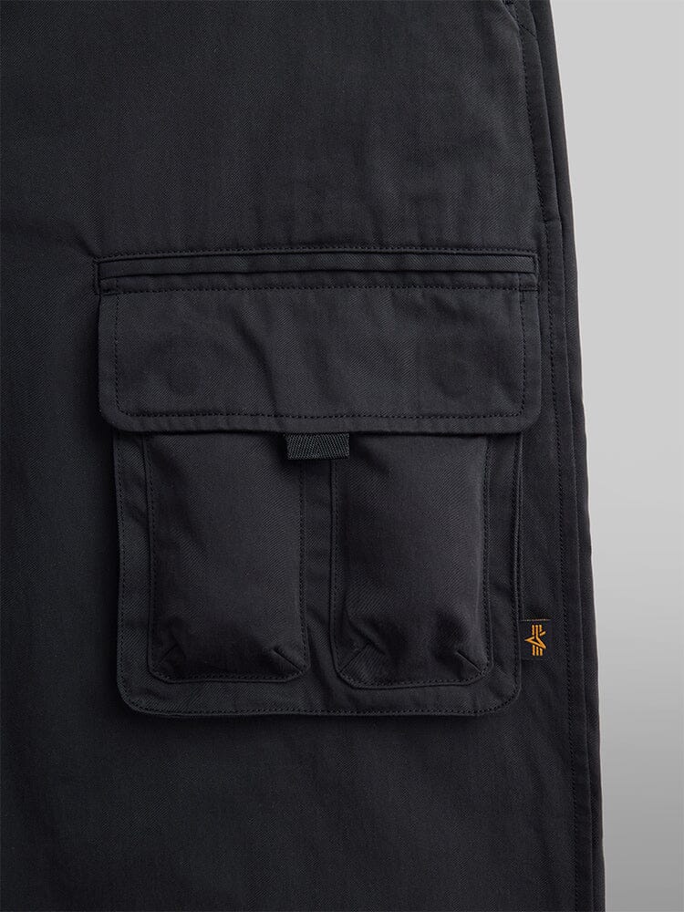 Close-up of a black cargo pocket on dark fabric with an elastic waistband, featuring visible stitching and a small Alpha Industries logo on the side. The product is the Oversized Tactical Pant.