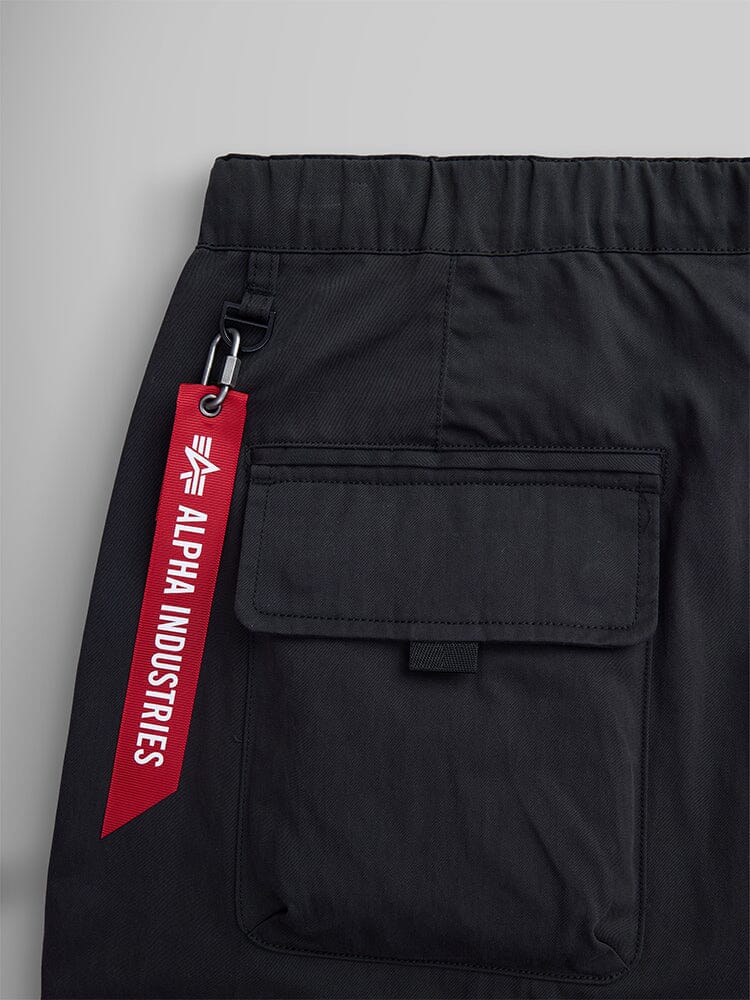 Close-up of black Oversized Tactical Pant by Alpha Industries, showcasing a flattering flap pocket and a striking red brand tag attached to the elastic waistband.