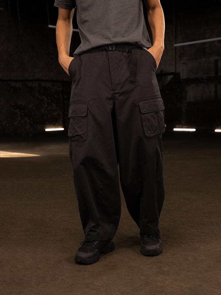 A person wearing an oversized gray shirt and the Oversized Tactical Pant by Alpha Industries, featuring baggy black cargo-style with an elastic waistband, stands indoors with hands casually tucked into the deep pockets.