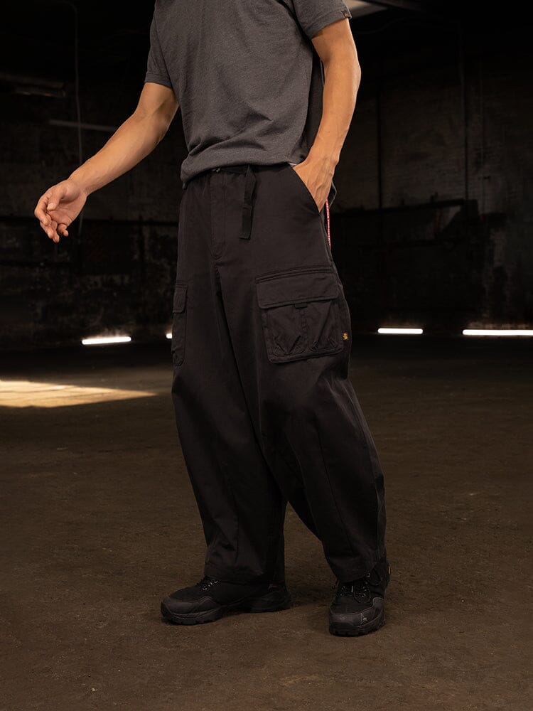 In a dimly lit room, the person sports a gray t-shirt and pairs it with Alpha Industries' Oversized Tactical Pant, which features an elastic waistband and ample cargo pockets, complemented by black shoes.