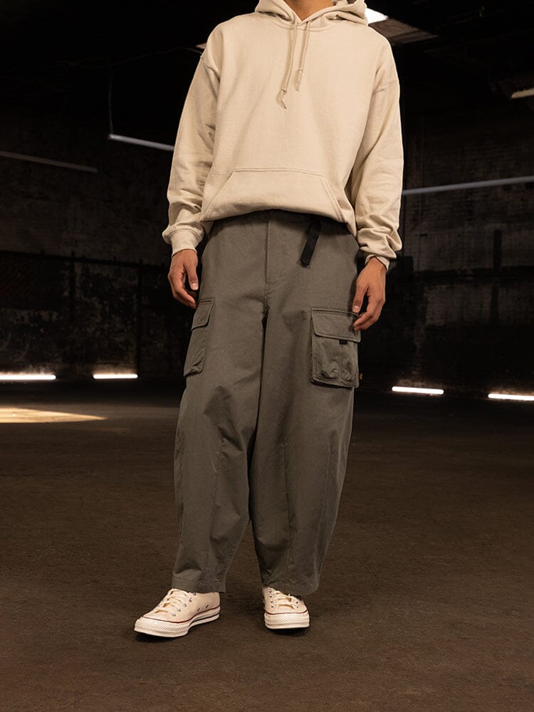 A person wearing a beige hoodie, Alpha Industries Oversized Tactical Pant featuring an elastic waistband, and white sneakers stands in a dimly lit room.