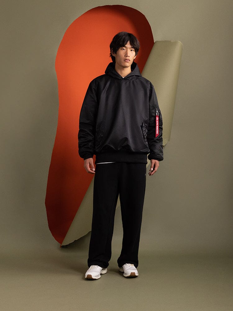 A person wearing a black Pull-Over MA-1 Jacket Hoodie from Alpha Industries stands clad in black pants and hoodie with the iconic flight nylon style complete with utility pockets.