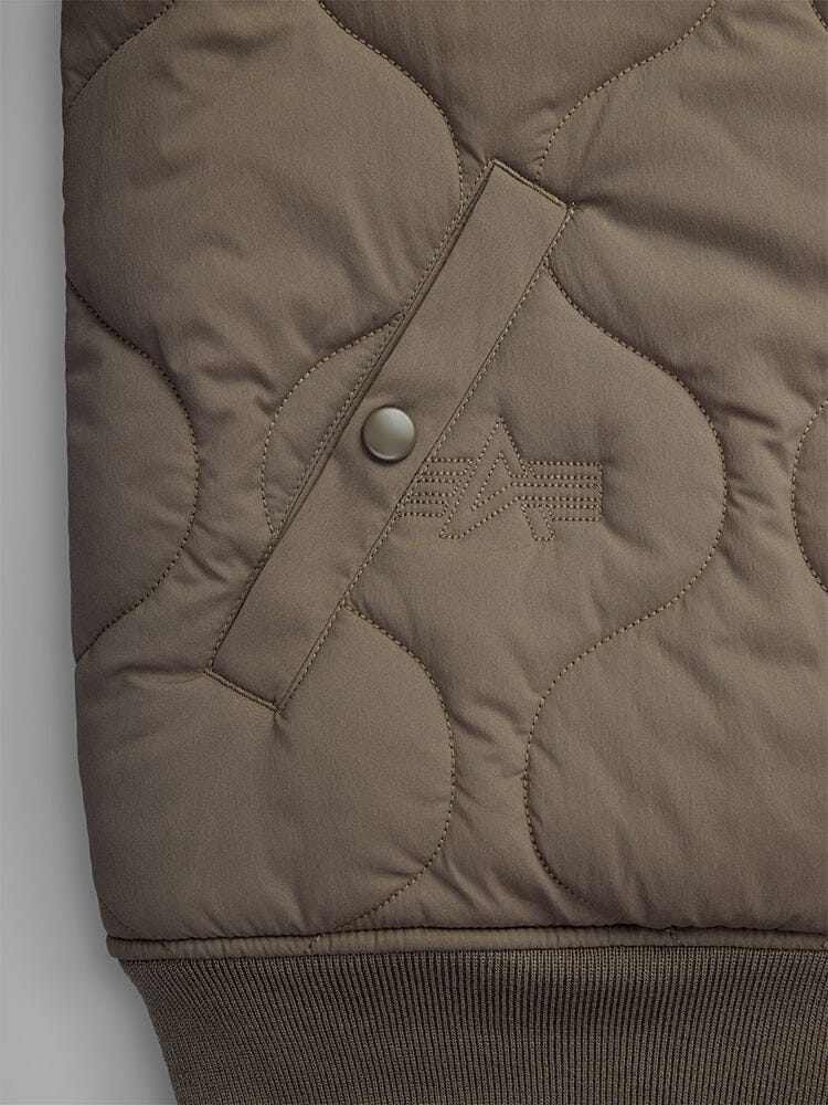 Close-up of a pocket from the Reversible Onion Quilted MA-1 Bomber Jacket by Alpha Industries, showcasing an olive-green quilted design with a stitched emblem and buttoned strap detail.
