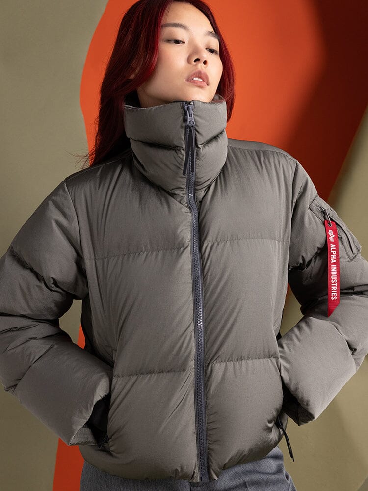 A person wearing the Alpha Industries Sierra Short Puffer W, a gray water-resistant parka with a distinctive red tag on the sleeve.
