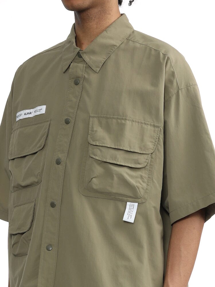 Unfrm Nylon Utility Shirt by Alpha Industries, olive green short-sleeve button-up with patch pockets, visible white tags, and front button closures.