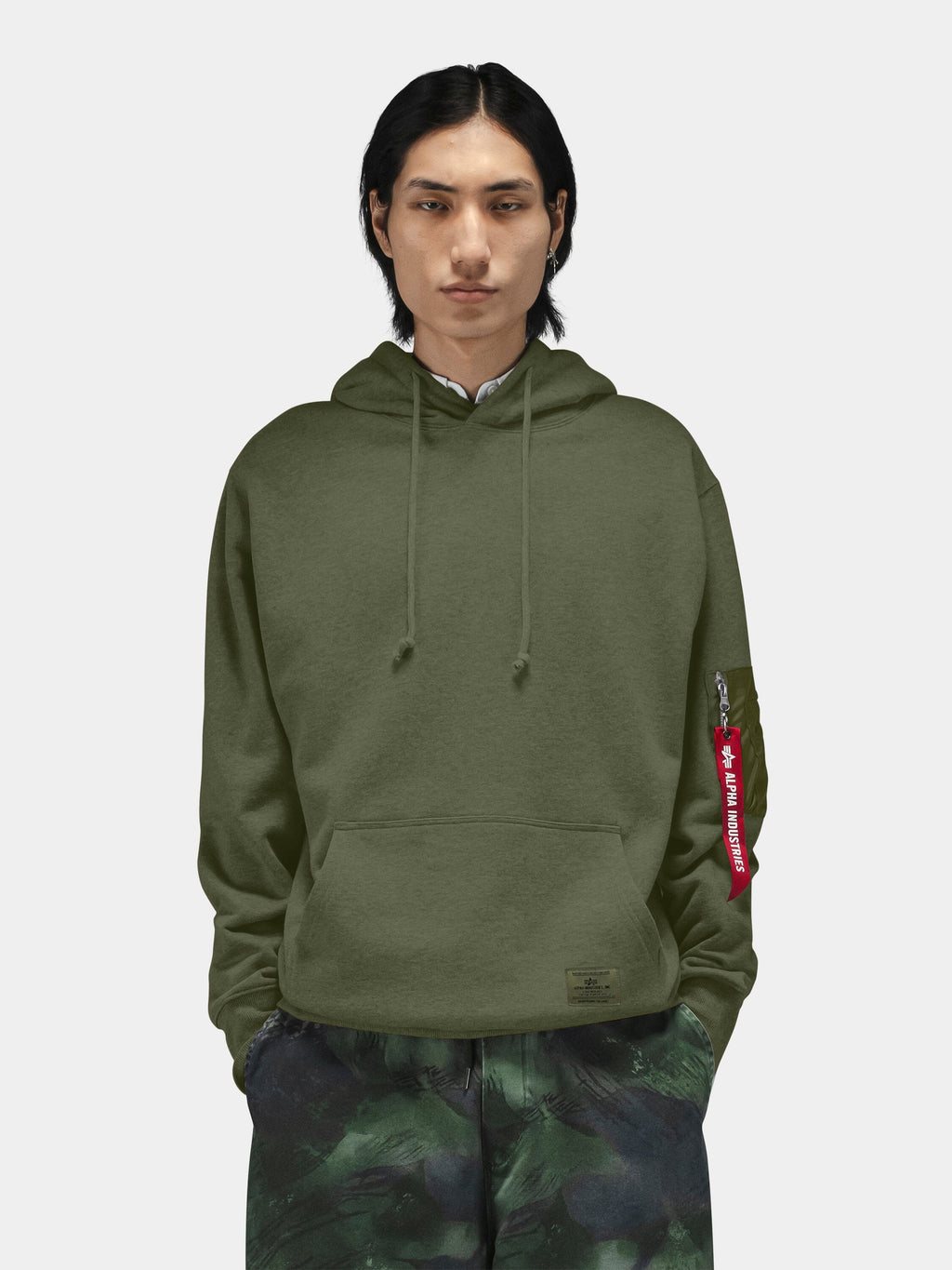 Alpha Industries Utility Pocket Hoodie with olive green MA-1 utility sleeve pocket, paired with camouflage pants.
