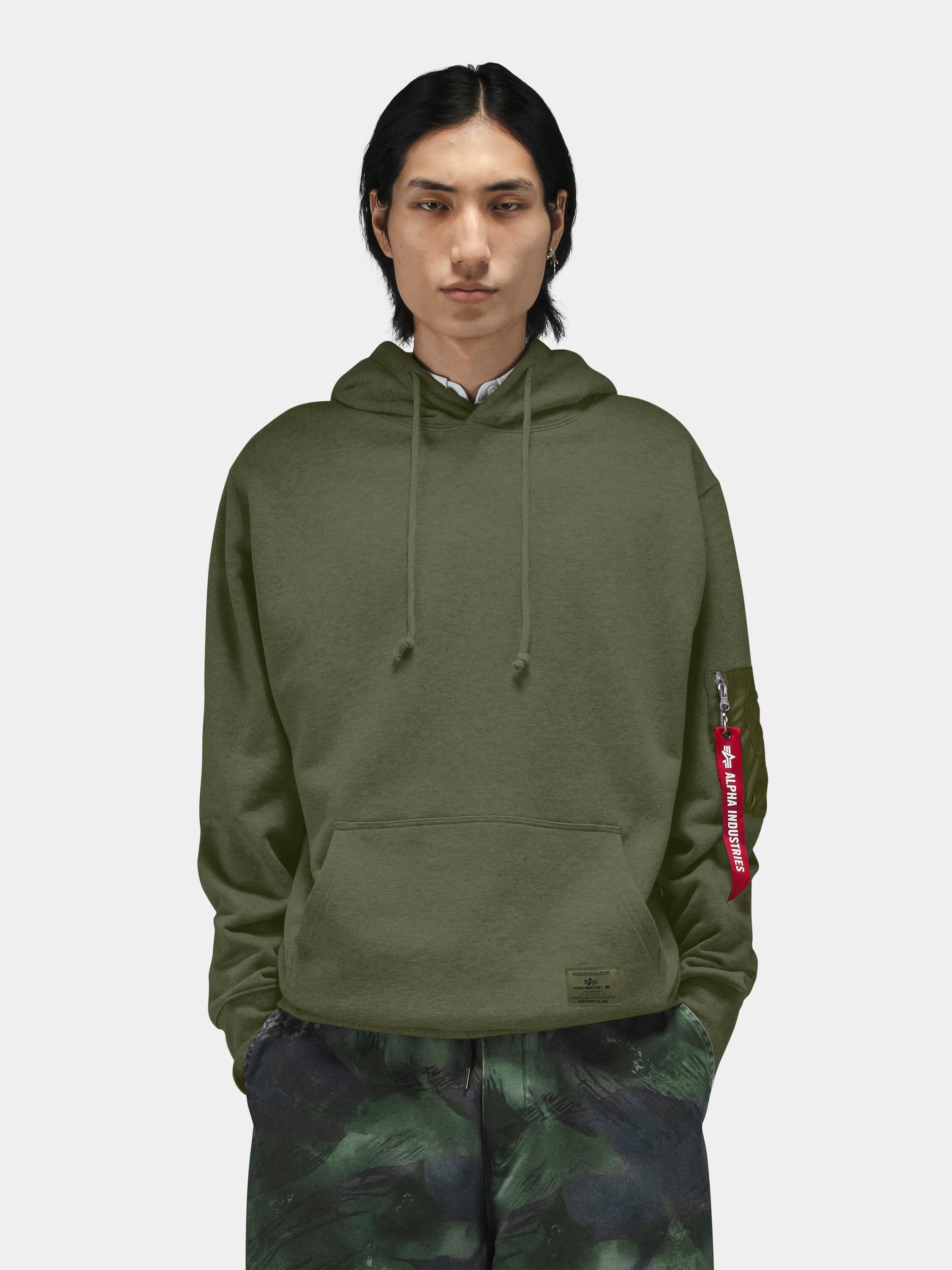 Alpha Industries Utility Pocket Hoodie with olive green MA-1 utility sleeve pocket, paired with camouflage pants.