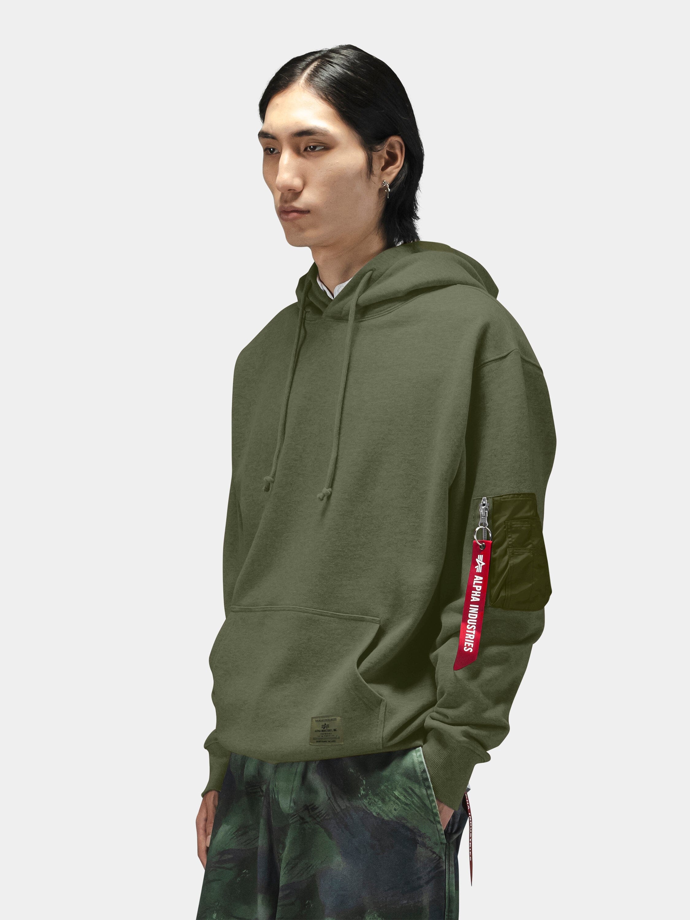 Alpha Industries Utility Pocket Hoodie in olive green with red Remove Before Flight tag on MA-1 sleeve pocket and green camo pants.