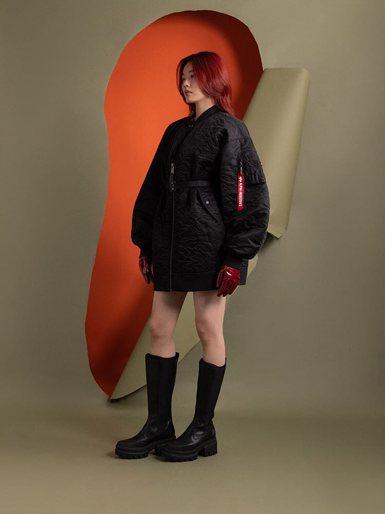 A person standing to the side wearing the Women's Camo Quilted Belted MA-1 Bomber Jacket in black by Alpha Industries.