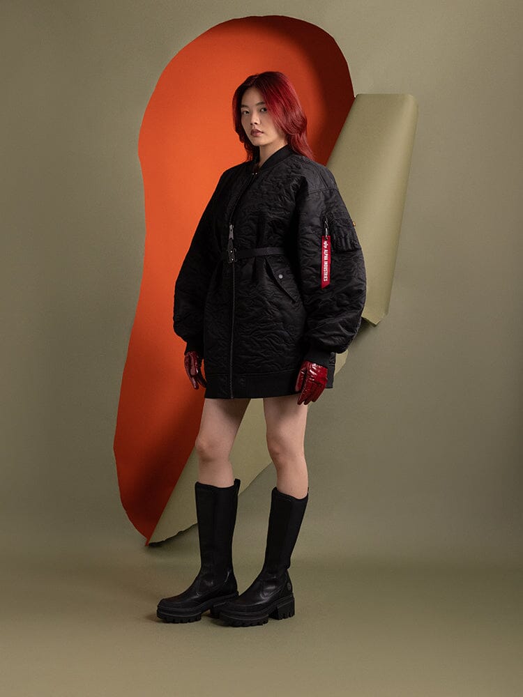 A person wearing the Women's Camo Quilted Belted MA-1 Bomber Jacket in black from Alpha Industries, along with red gloves and black boots.