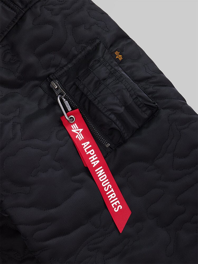 Close-up of the Women's Camo Quilted Belted MA-1 Bomber Jacket in black from Alpha Industries, featuring a red Alpha Industries tag attached to a zippered pocket. The fabric boasts quilted detailing, adding a subtle textured pattern.