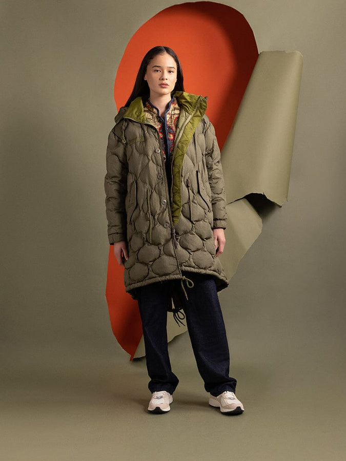 A person is wearing the Women's M-65 Fishtail Puffer in OG-107 green by Alpha Industries, featuring a long, onion-quilted design with a hood.