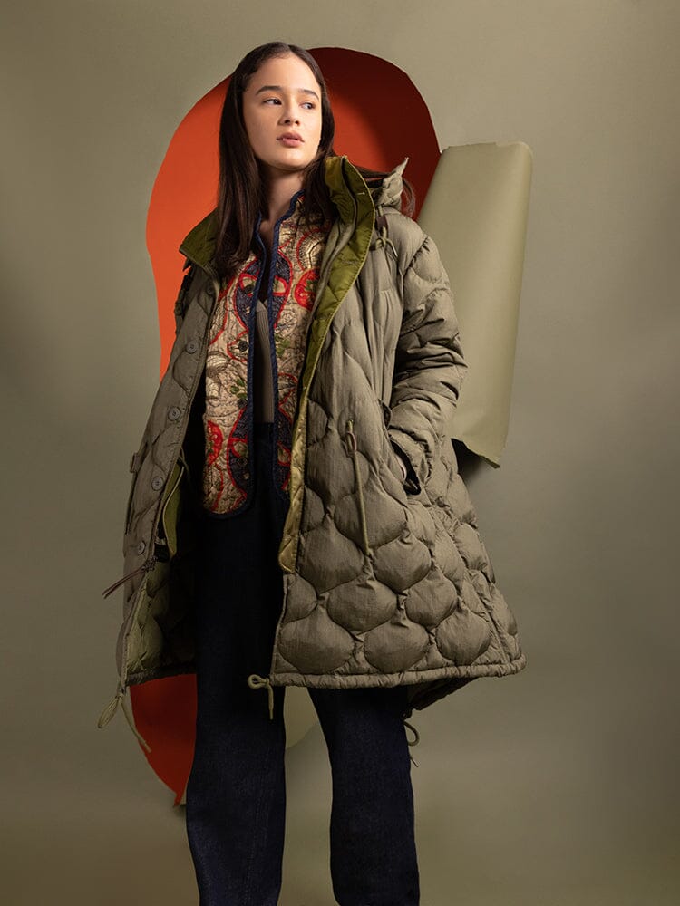 A person wearing the Women's M-65 Fishtail Puffer in green from Alpha Industries and a patterned sweater stands.