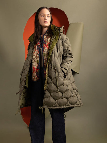 A person wearing the Women's M-65 Fishtail Puffer in green from Alpha Industries and a patterned sweater stands.