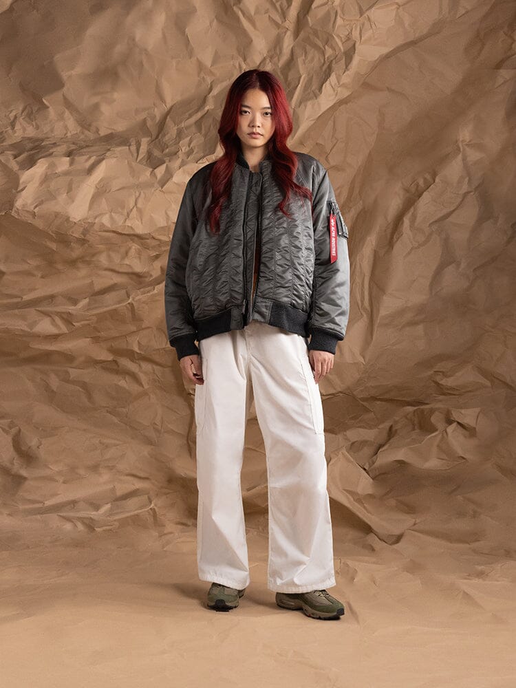 A person wearing the Women's MA-1 Shirring Bomber Jacket in field gray by Alpha Industries, along with white pants and dark shoes.