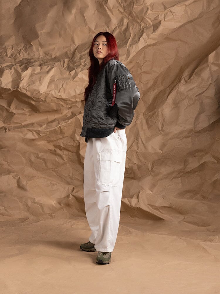 Side-view of a person wearing the Women's MA-1 Shirring Bomber Jacket by Alpha Industries in gray, paired with white cargo pants featuring a utility pocket.