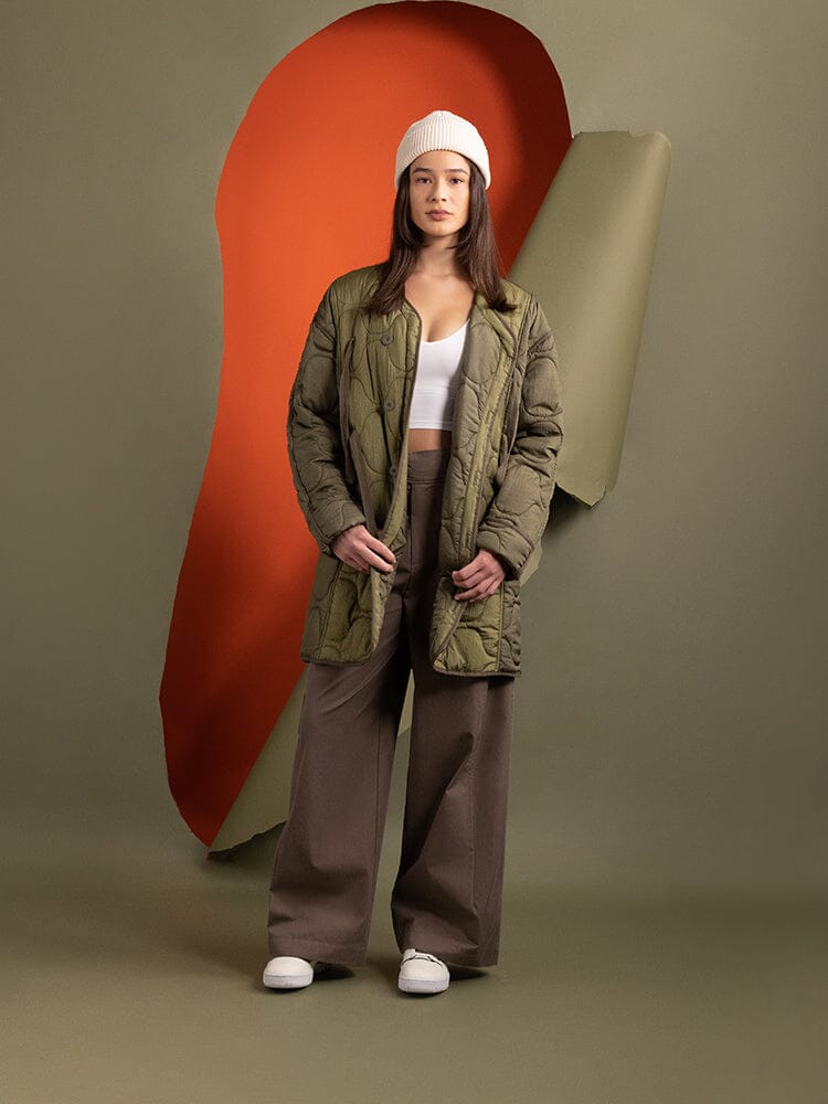 A person stands confidently, showcasing the layering trend, wearing Alpha Industries Women's Mixed Media Quilted Long Liner in OG-107 Green, and a white top.