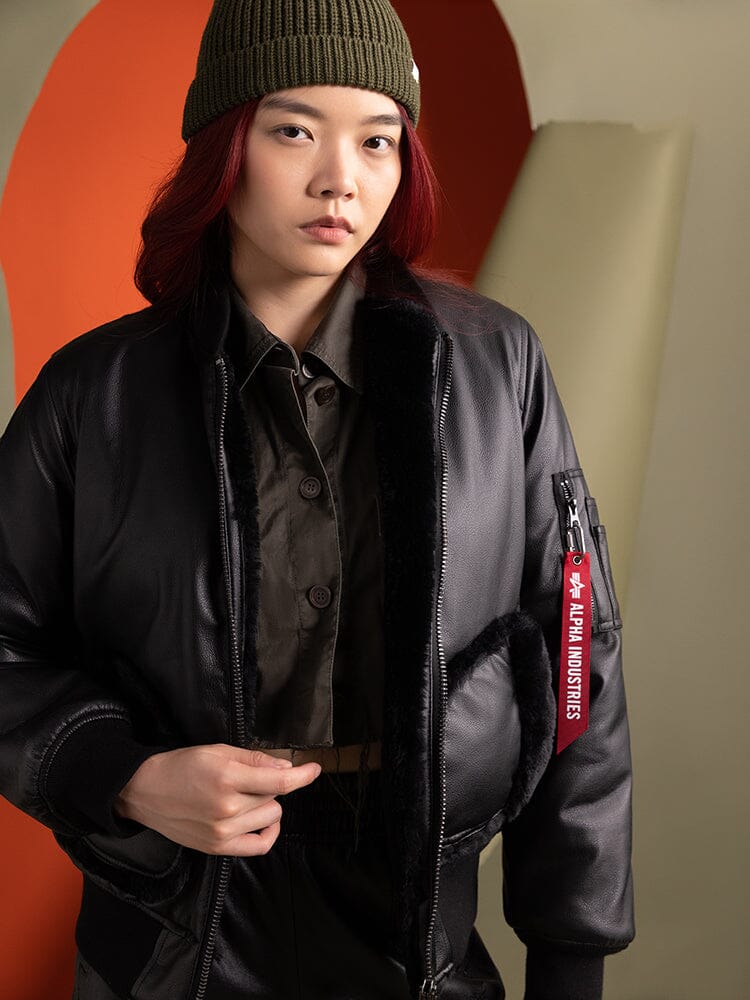 A person wearing the Women's Reversible Faux Leather MA-1 Bomber Jacket in black by Alpha Industries and a green beanie stands confidently.
