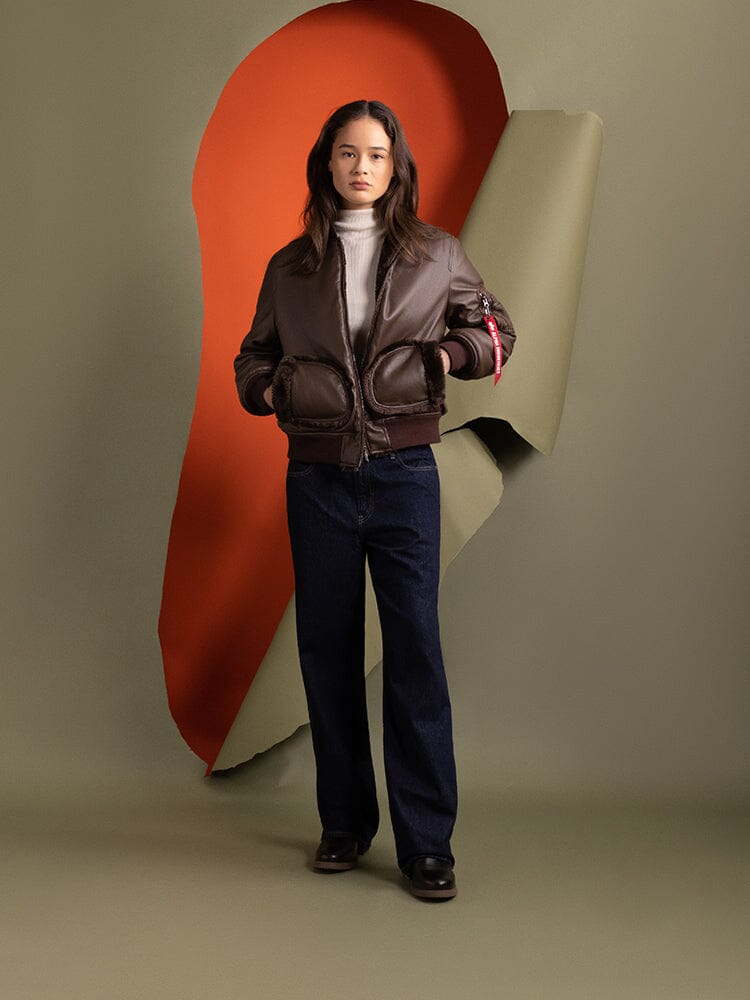 A person stands wearing the Alpha Industries Women's Reversible Faux Leather MA-1 Bomber Jacket in brown chocolate paired with jeans.