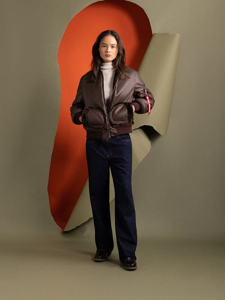 A person stands wearing the Alpha Industries Women's Reversible Faux Leather MA-1 Bomber Jacket in brown chocolate paired with jeans.