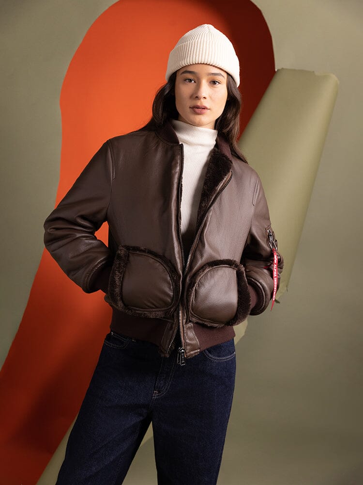 A person wearing the Women's Reversible Faux Leather MA-1 Bomber Jacket in brown chocolate by Alpha Industries, paired with a white beanie. 