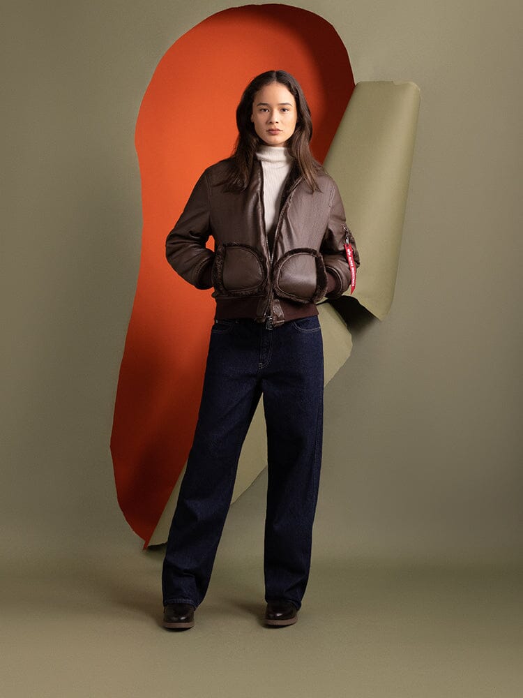 A person stands wearing an Alpha Industries Women's Reversible Faux Leather MA-1 Bomber Jacket in brown chocolate, a white turtleneck, and dark pants.