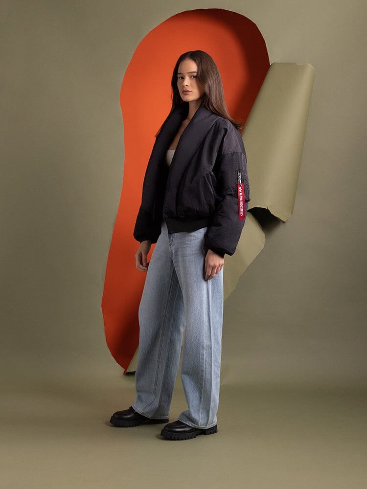 A person standing to the side wearing the Women's Shawl Collar Bomber Jacket in black by Alpha Industries.