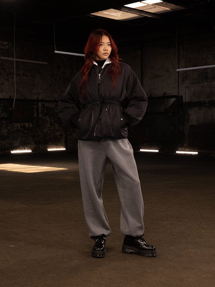 An individual with long hair wears the Women's Short Cinched Waist Jacket by Alpha Industries, complemented by gray pants and black shoes.