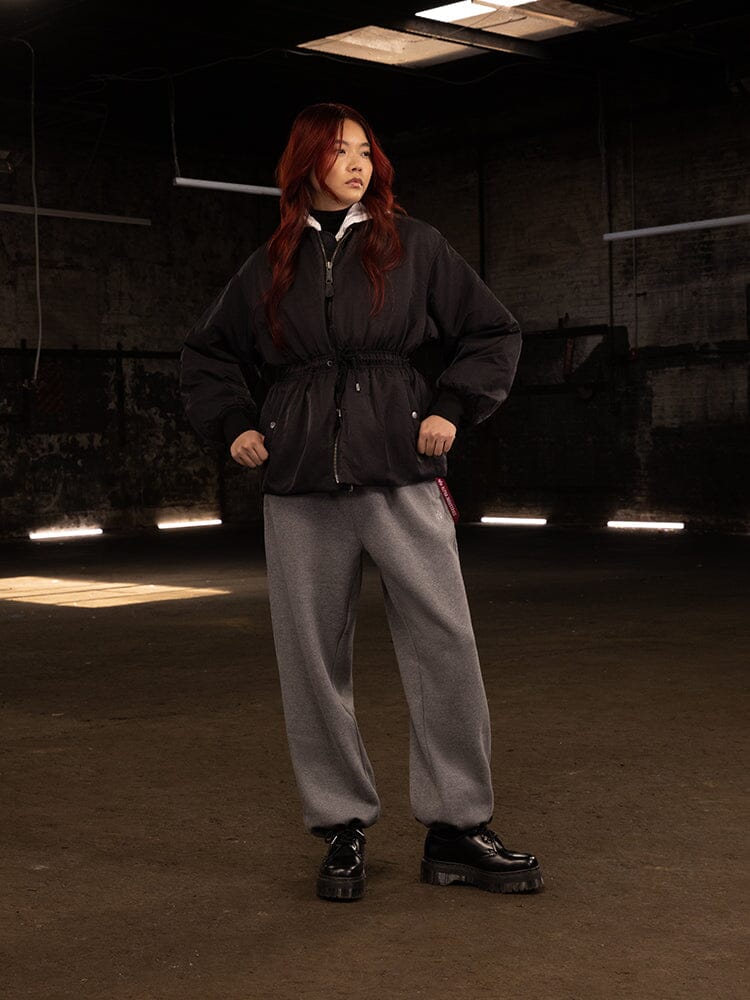 A person with long red hair stands confidently in the Women's Short Cinched Waist Jacket by Alpha Industries, featuring quilted lining and an adjustable drawcord at the waist. They complete the look with gray pants and black shoes.