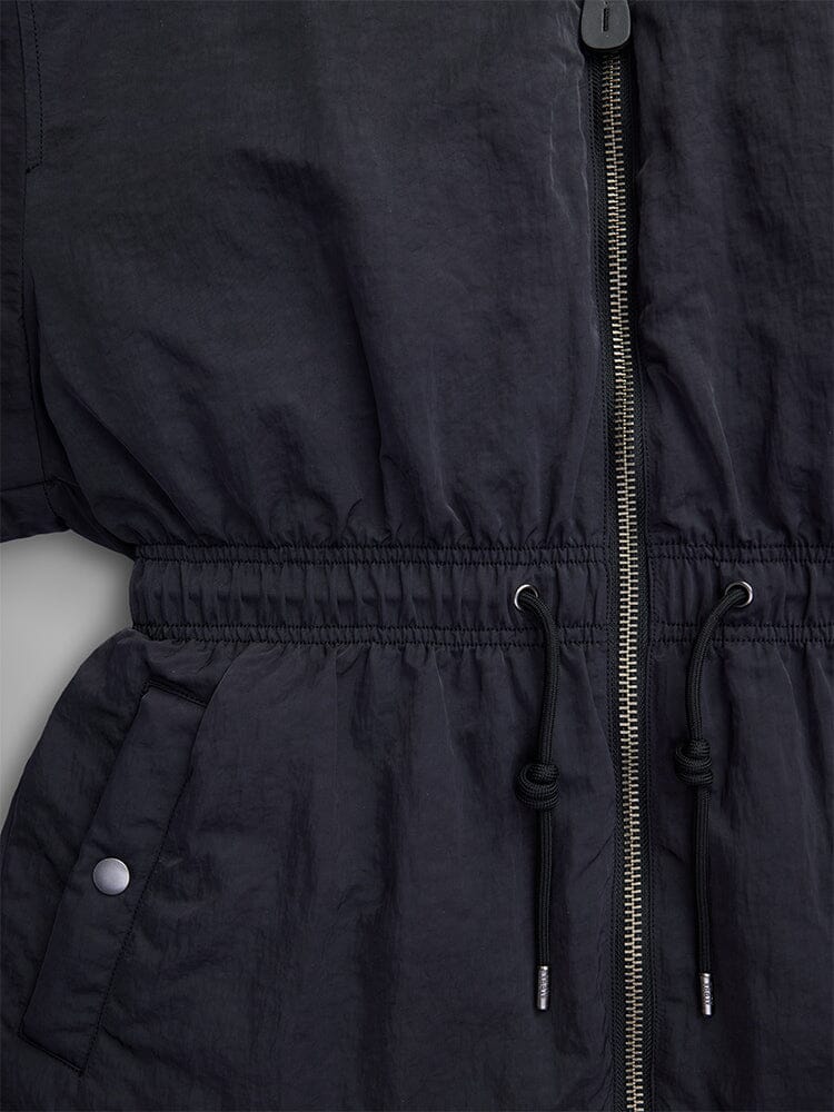 Close-up of the Alpha Industries Women's Short Cinched Waist Jacket in black nylon, showcasing a zipper, adjustable drawcord waist, and welt pockets. The design is enhanced with buttoned details and a cozy quilted lining.