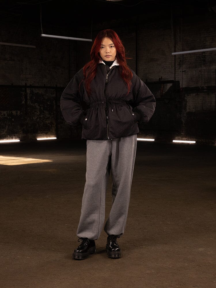 A person with long red hair stands wearing the Women's Short Cinched Waist Jacket by Alpha Industries and gray pants.