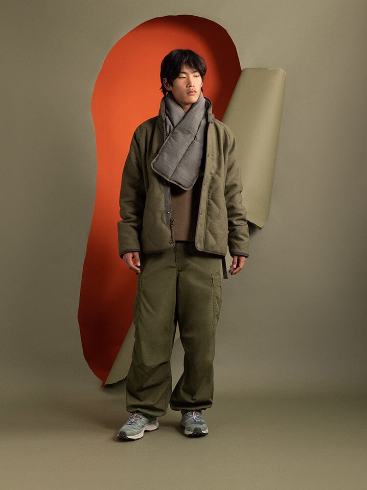 A person in a green outfit and gray scarf stands, dressed in a stylish Wool Deck Jacket from Alpha Industries.