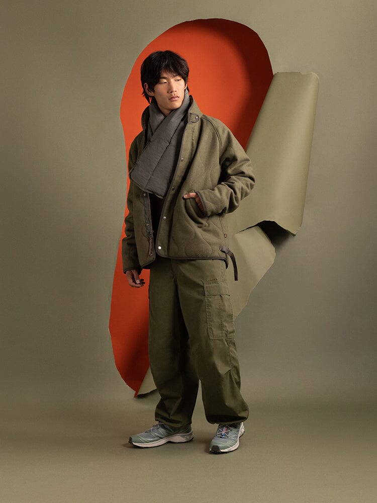 A person dressed in an Alpha Industries olive green Wool Deck Jacket, paired with a scarf, stands.