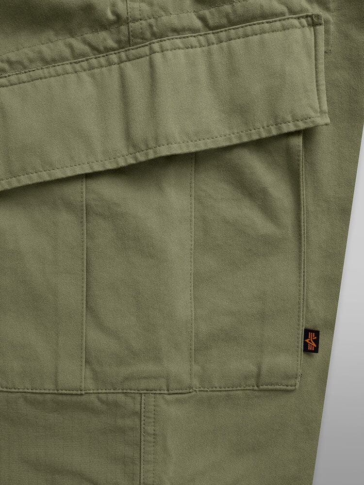 Alpha Industries Acu Pant in olive green with visible stitching, flap utility pocket, nylon knee patches, and small black and orange rectangular tag on the seam.