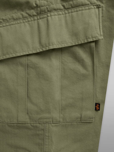 Alpha Industries Acu Pant in olive green with visible stitching, flap utility pocket, nylon knee patches, and small black and orange rectangular tag on the seam.