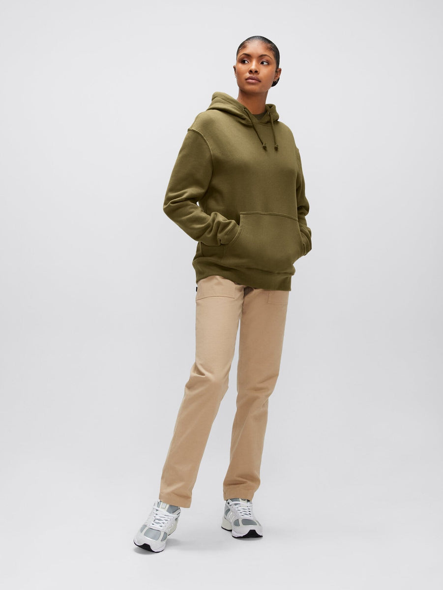 A person wearing the Alpha Hoodie in olive green from Alpha Industries, Inc., made from soft cotton fabric, pairs it stylishly with beige pants and white sneakers.