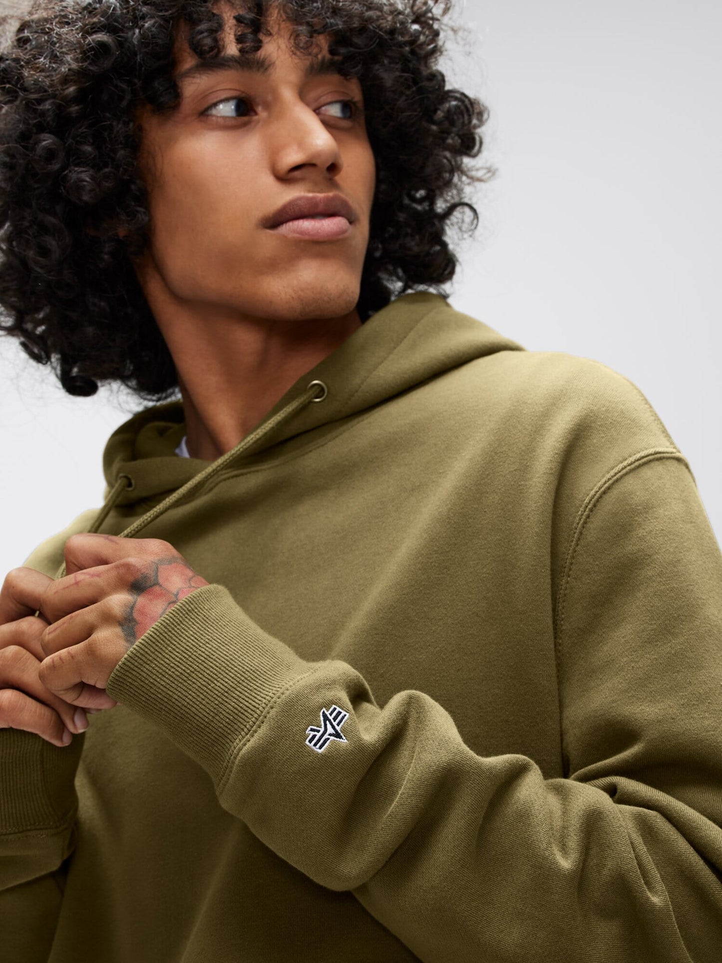 A person wearing an Alpha Hoodie by Alpha Industries, Inc. is donning a green unisex hoodie made from soft cotton fabric with an Alpha applique logo on the sleeve. They are looking to the side with hands clasped, and their curly hair frames a neutral expression.