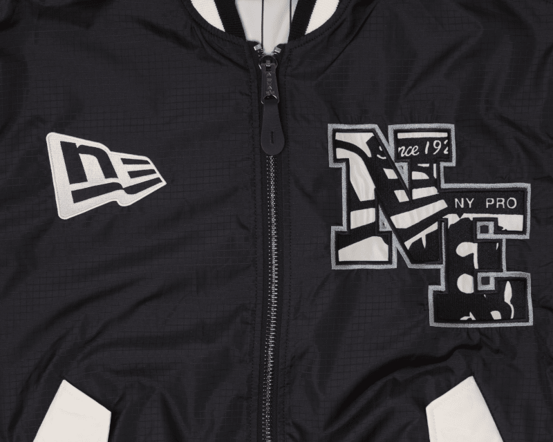 The Alpha x New Era L-2B Bomber Jacket by Alpha Industries showcases a close-up of its black ripstop fabric adorned with embroidered logos, including NE and NY Pro in white and grey. This bomber jacket features a front zipper along with black-and-white striped accents.