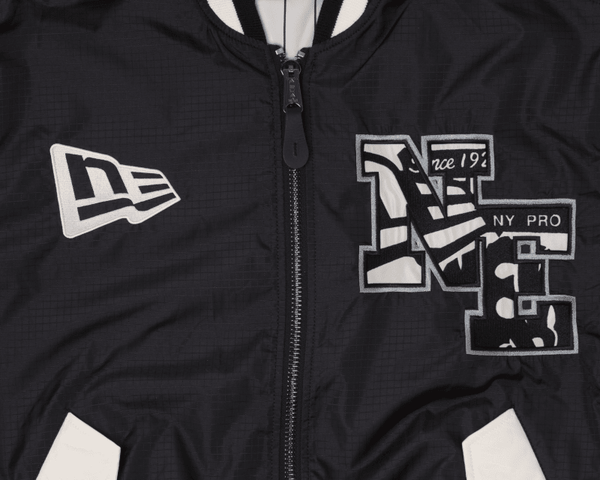 The Alpha x New Era L-2B Bomber Jacket by Alpha Industries showcases a close-up of its black ripstop fabric adorned with embroidered logos, including NE and NY Pro in white and grey. This bomber jacket features a front zipper along with black-and-white striped accents.