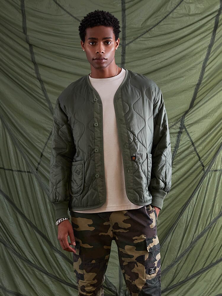 Alpha Industries Als/92 Liner in olive, worn over beige t-shirt and camouflage pants.