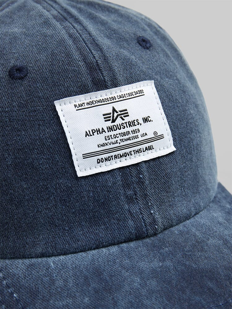 A close-up of the Battlewash Cap in blue denim highlights a white label with black text from Alpha Industries. The vintage-inspired style includes manufacturing details and a warning to not remove the label.