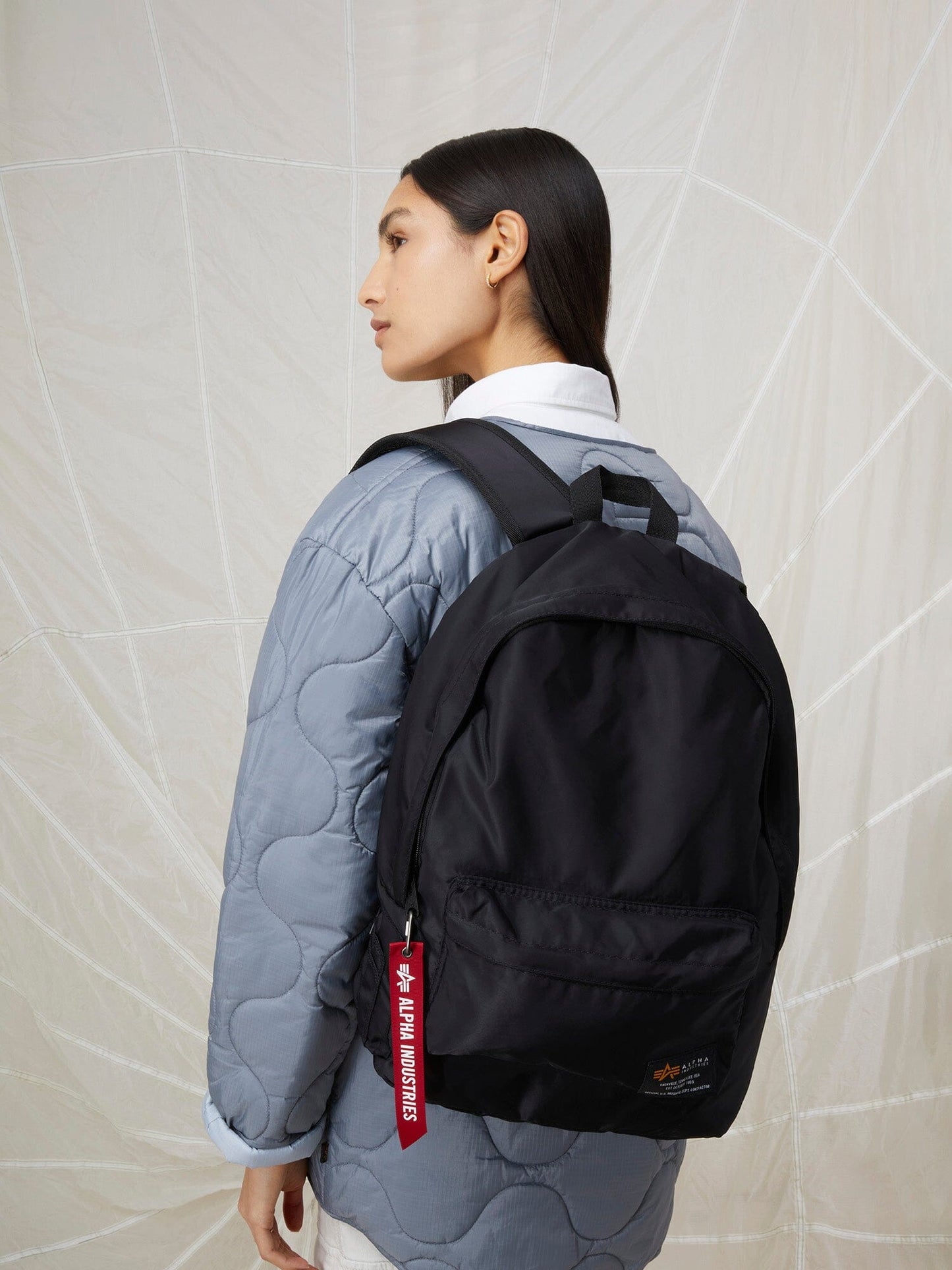A women carries the ultra-lightweight Crew Backpack by Alpha Industries showcasing a red tag and adjustable nylon straps.